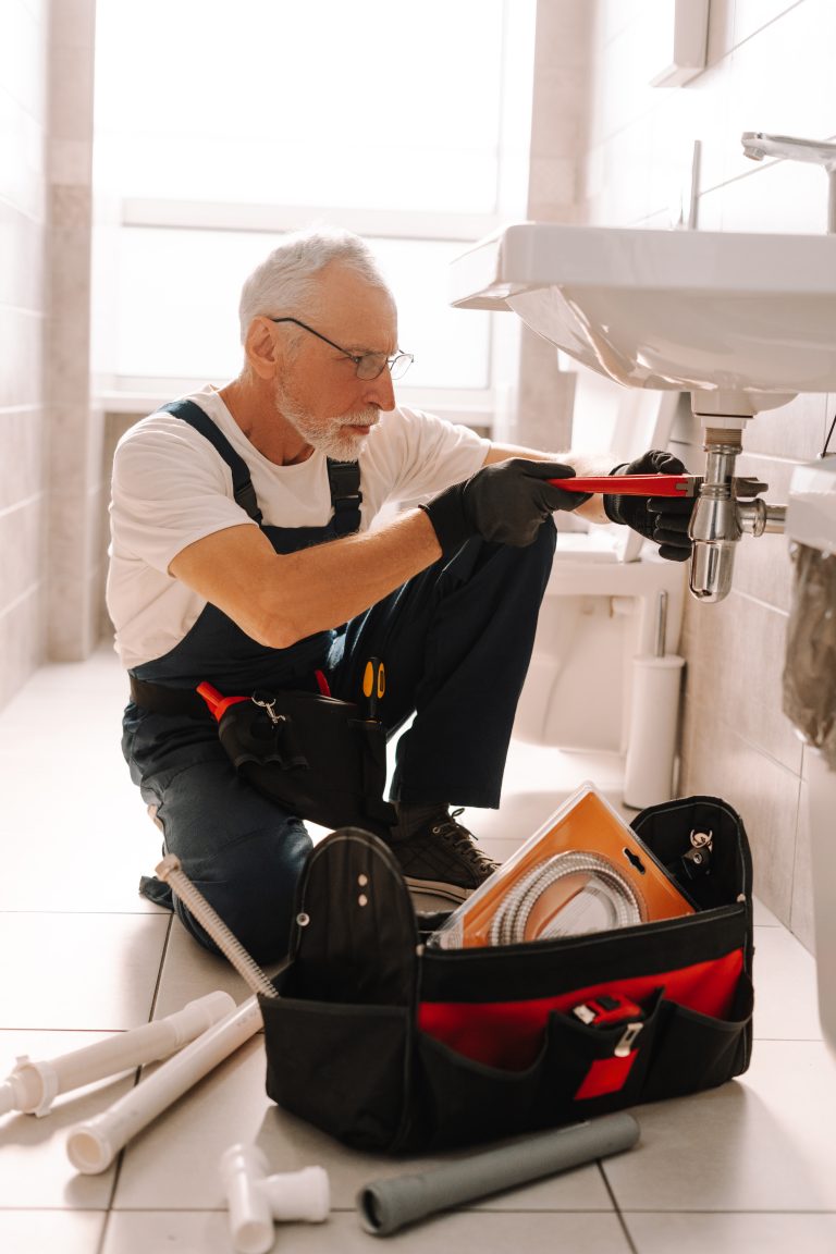 Senior plumber wearing blue overalls is installing a sink using an adjustable wrench in a bathroom. His tool bag is open on the floor next to him